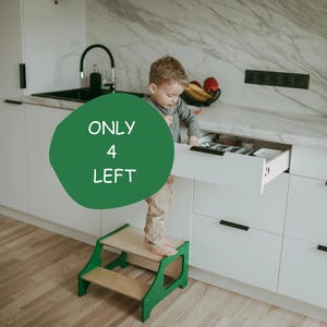 May include: A green and natural wood step stool with a child standing on it, reaching into a white kitchen drawer. The kitchen has white cabinets and a marble countertop. Text on a green circle reads "ONLY 4 LEFT".