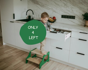 Puede incluir: Un taburete verde y de madera natural con un niño de pie sobre él, alcanzando un cajón de cocina blanco. La cocina tiene gabinetes blancos y una encimera de mármol. El texto en un círculo verde dice "ONLY 4 LEFT".