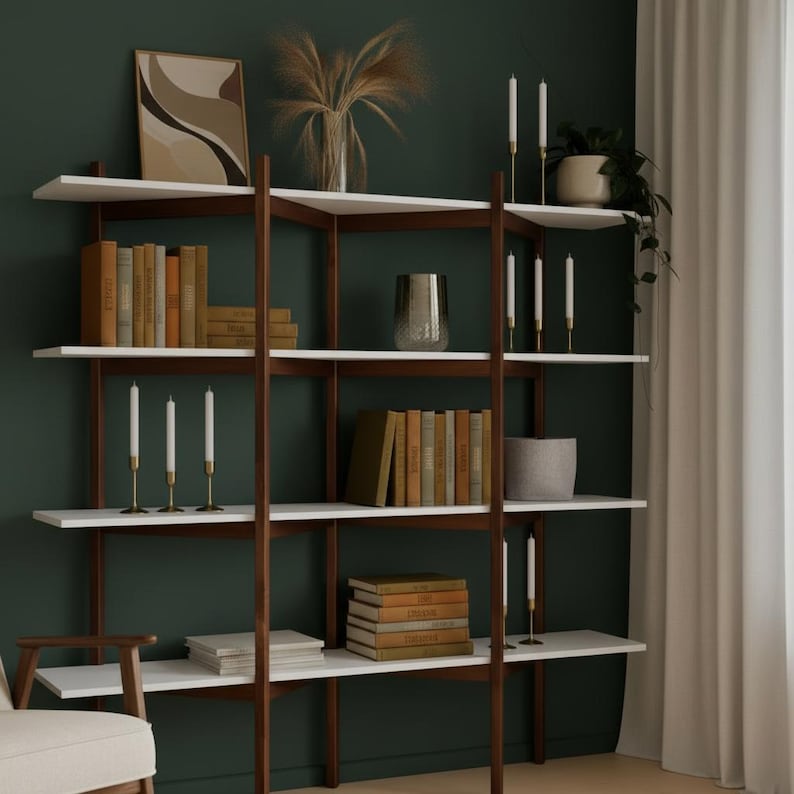 May include: A modern wooden bookshelf with white shelves and dark brown supports. The shelves are filled with books, candles, and decorative items. The background is a dark green wall, and a white curtain is visible on the right.