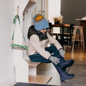 May include: A white wooden entryway organizer with a shelf, hooks, and a small step stool. A child wearing a blue hat with a yellow smiley face is sitting on the step stool and putting on blue rain boots.