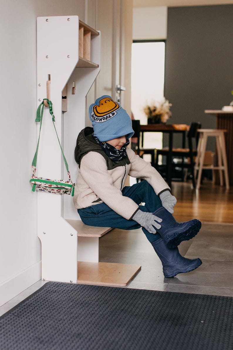May include: A white wooden entryway organizer with a shelf, hooks, and a small step stool. A child wearing a blue hat with a yellow smiley face is sitting on the step stool and putting on blue rain boots.