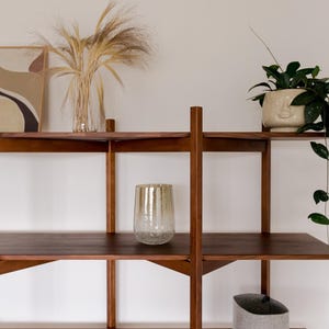 May include: A wooden shelving unit with three shelves, holding decorative items. A clear glass vase with dried plants, a textured glass, a potted plant, and a gray felt container are displayed. The shelves are a dark brown color.