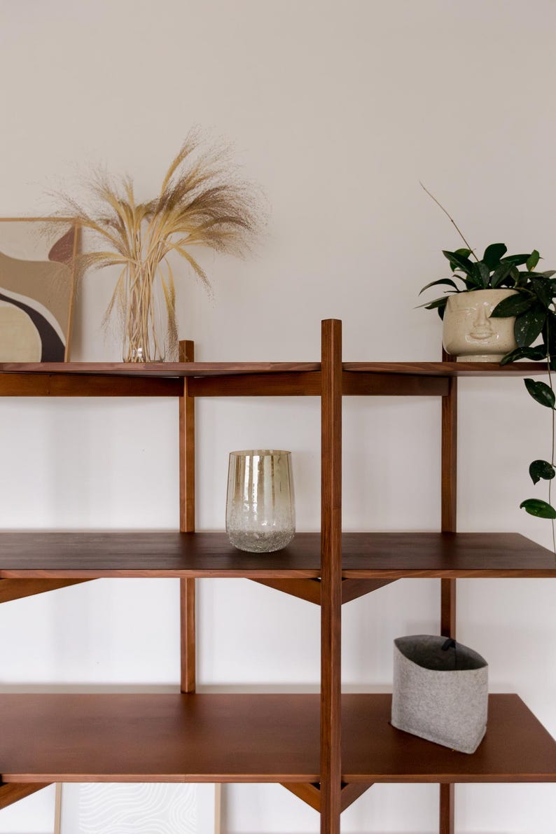 May include: A wooden shelving unit with three shelves, holding decorative items. A clear glass vase with dried plants, a textured glass, a potted plant, and a gray felt container are displayed. The shelves are a dark brown color.