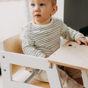 May include: A white wooden high chair with a light brown wooden table top. A young child is sitting in the chair wearing a white and black striped long-sleeved shirt and beige pants.