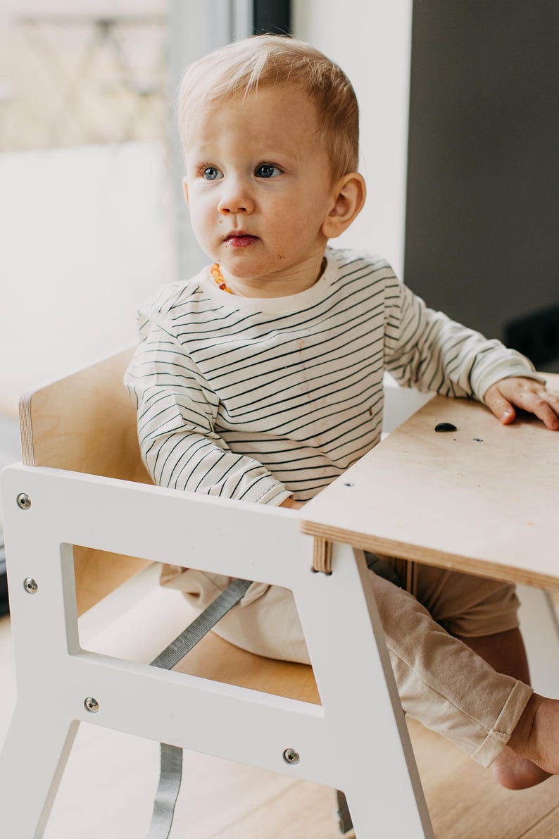 May include: A white wooden high chair with a light brown wooden table top. A young child is sitting in the chair wearing a white and black striped long-sleeved shirt and beige pants.