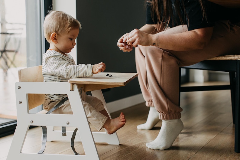 May include: A white wooden high chair with a light wood tray and a safety harness. A young child sits in the chair, while an adult sits on a black chair in the background.