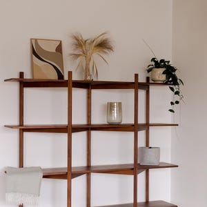 May include: A wooden bookshelf with multiple shelves displays decorative items. A framed abstract art piece, a vase with dried plants, a potted plant, a textured glass vase, a small gray pot, and a folded blanket are visible.
