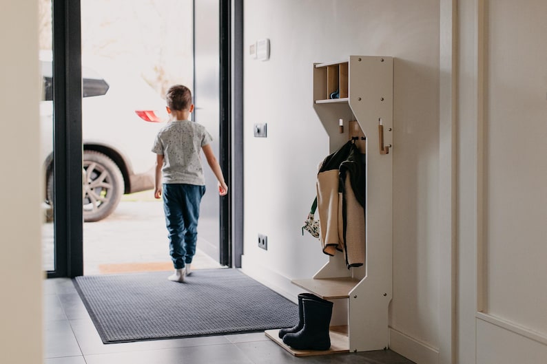 May include: A white wooden coat rack with a shelf, hooks, and a shoe storage area. A child is walking through a doorway in the background.