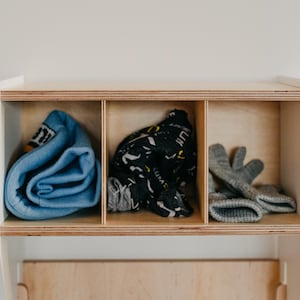 May include: A white wooden shelf with three compartments. The compartments contain a blue rolled up sweater, a black and white patterned scarf, and a pair of gray mittens.