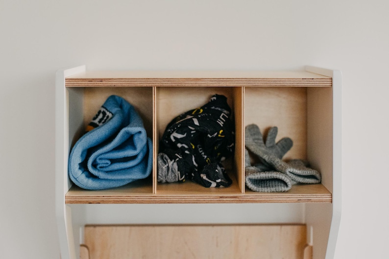 May include: A white wooden shelf with three compartments. The compartments contain a blue rolled up sweater, a black and white patterned scarf, and a pair of gray mittens.