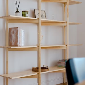 May include: A light-colored wooden bookshelf with multiple shelves. The shelves hold various decorative items, including books, candles, and small framed pictures. The bookshelf is against a white wall.