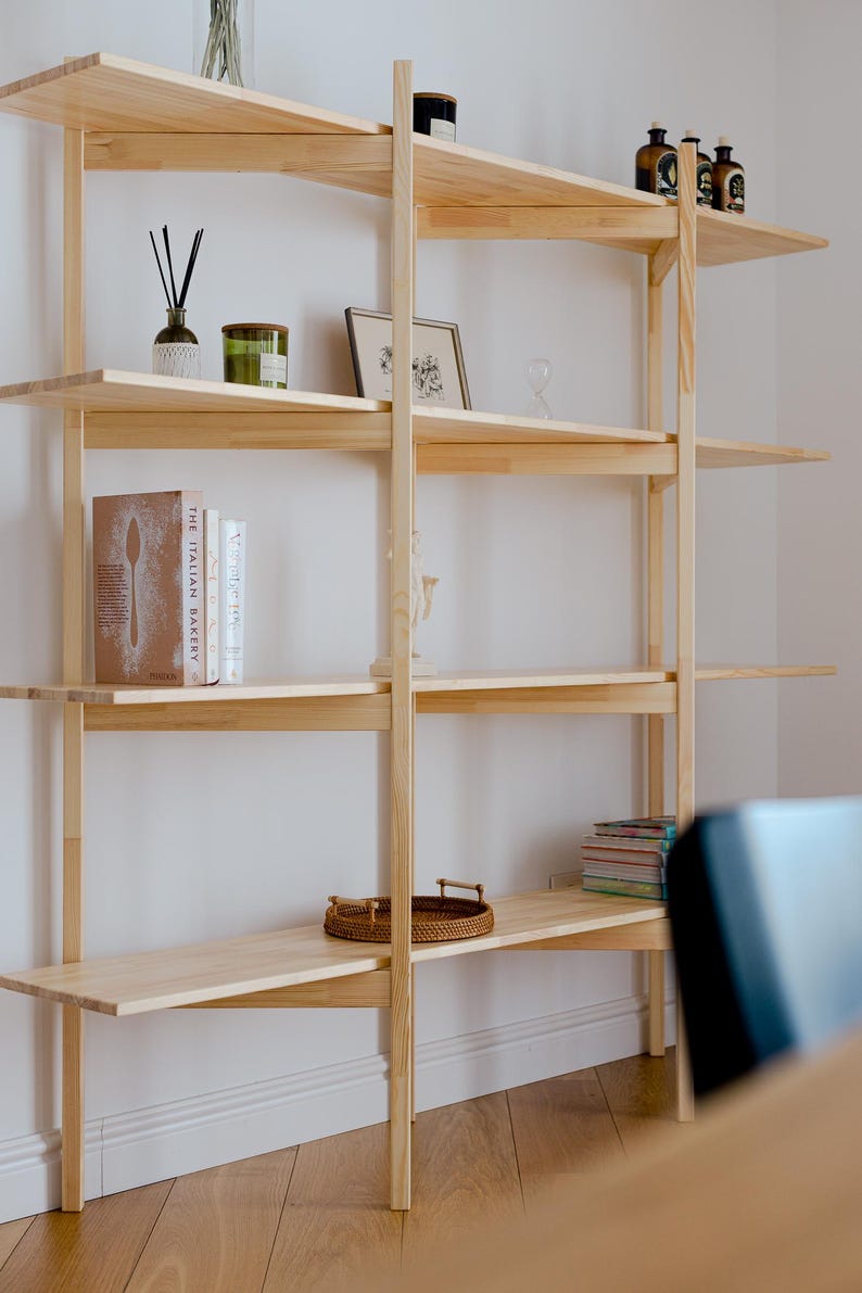 May include: A light-colored wooden bookshelf with multiple shelves. The shelves hold various decorative items, including books, candles, and small framed pictures. The bookshelf is against a white wall.