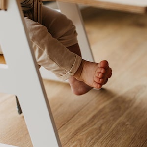 May include: A white wooden high chair with a child's bare feet dangling over the side. The child is wearing beige pants and is sitting at a table.