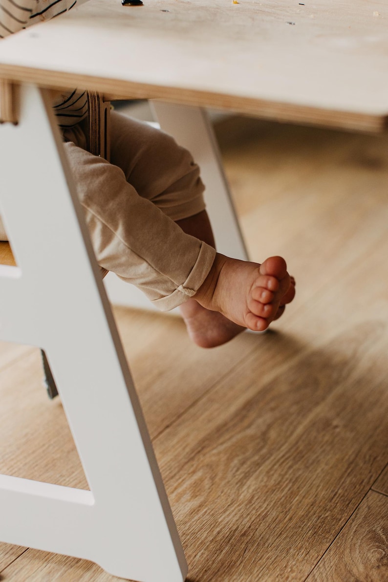 May include: A white wooden high chair with a child's bare feet dangling over the side. The child is wearing beige pants and is sitting at a table.