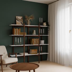 May include: A modern bookshelf with white shelves and dark wood supports. The shelves are filled with books, candles, and decorative items. A round wooden coffee table and a cream-colored armchair are in the foreground. The background is a dark green wall.