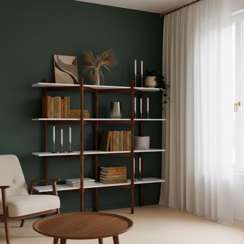 May include: A modern bookshelf with white shelves and dark wood supports. The shelves are filled with books, candles, and decorative items. A round wooden coffee table and a cream-colored armchair are in the foreground. The background is a dark green wall.