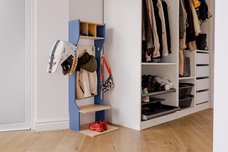 May include: A blue and wood entryway organizer with a helmet, jackets, and a purse hanging on it. Red shoes sit on the bottom shelf. A white closet with clothes and shoes is to the right.