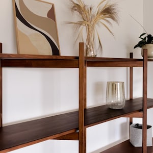 May include: A dark brown wooden wall shelf unit with three shelves. The shelves hold a framed abstract art piece, a glass vase with dried plants, a potted plant, a decorative glass, and a gray fabric container.