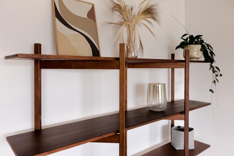 May include: A dark brown wooden wall shelf unit with three shelves. The shelves hold a framed abstract art piece, a glass vase with dried plants, a potted plant, a decorative glass, and a gray fabric container.