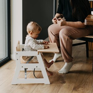 May include: A white wooden high chair with a light wood tabletop and a gray safety harness. A young child sits in the chair with a woman sitting nearby.