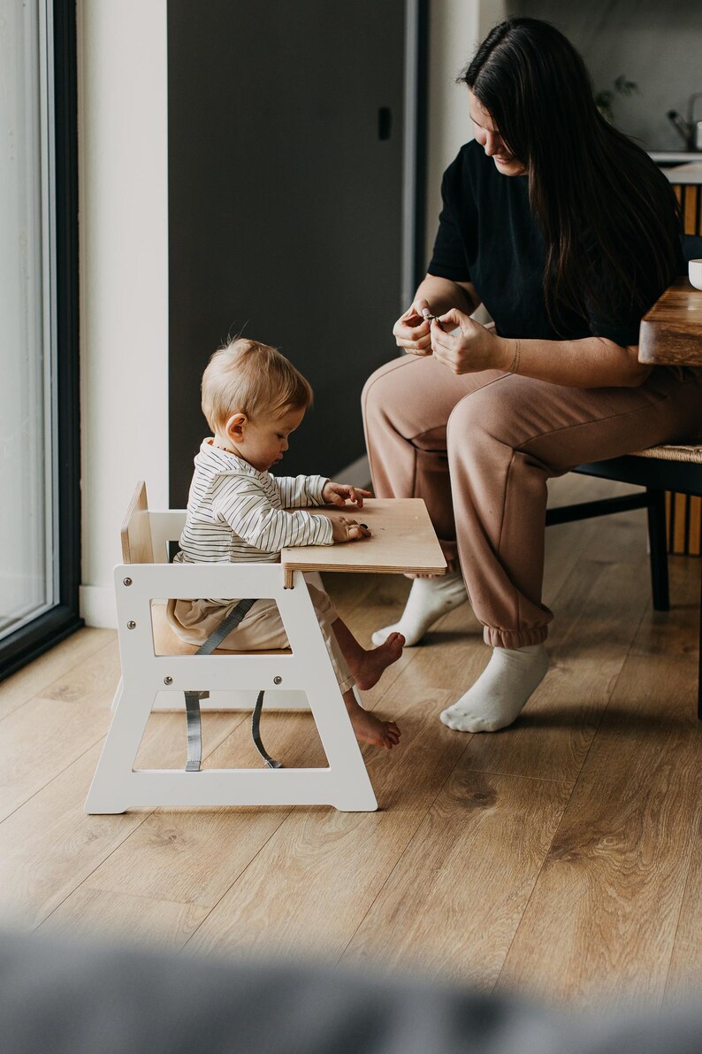 May include: A white wooden high chair with a light wood tabletop and a gray safety harness. A young child sits in the chair with a woman sitting nearby.