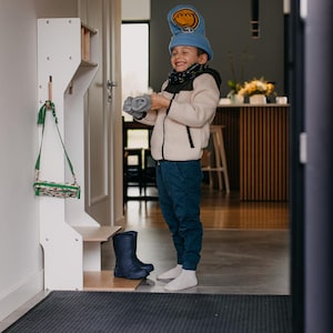 May include: A white wooden coat rack with a green and white bag hanging on a hook. A child wearing a blue hat with a smiley face and a white jacket is standing in front of the rack. The child is holding a pair of blue gloves. The child is wearing blue jeans and white socks.