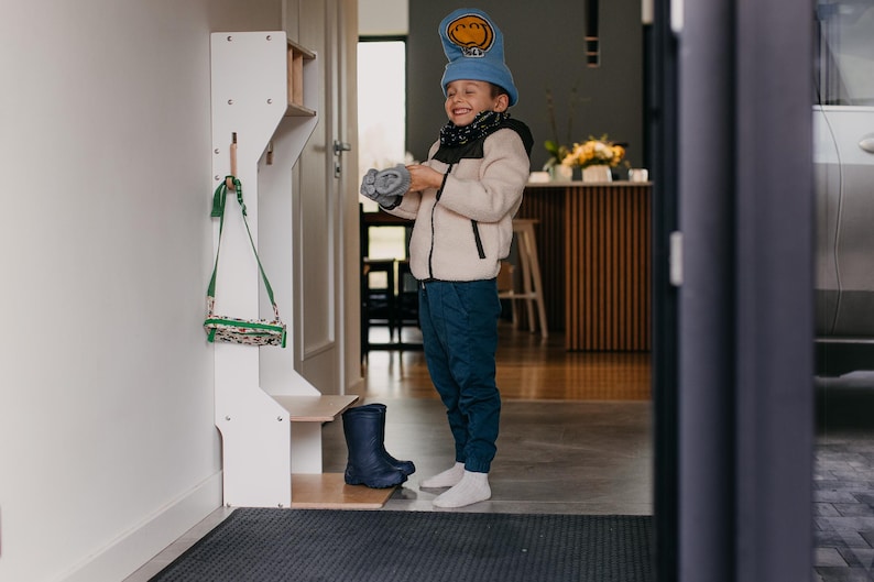 May include: A white wooden coat rack with a green and white bag hanging on a hook. A child wearing a blue hat with a smiley face and a white jacket is standing in front of the rack. The child is holding a pair of blue gloves. The child is wearing blue jeans and white socks.