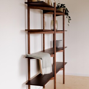 May include: A dark brown wooden bookshelf with four shelves. The shelves hold decorative items, including a framed artwork, a vase with dried plants, a potted plant, a gray basket, and a light green blanket with fringe.