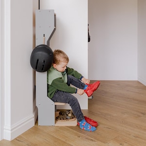 May include: A child is shown putting on a red shoe while seated on a gray wooden bench. A black helmet hangs on the side of the bench. The child is wearing a green and gray sweater, dark jeans, and blue socks with a pattern.