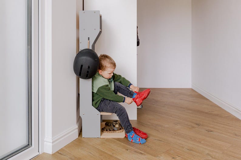 May include: A child is shown putting on a red shoe while seated on a gray wooden bench. A black helmet hangs on the side of the bench. The child is wearing a green and gray sweater, dark jeans, and blue socks with a pattern.