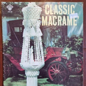 May include: A vintage book cover showing a white macrame hanging planter with tassels. The title "CLASSIC MACRAME" is displayed. A red antique car is in the background, with a green lawn and a house. The cover is on a brown surface.