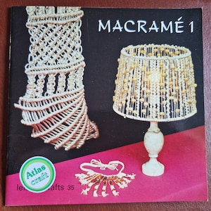 May include: The cover of a book titled "MACRAMÉ 1" showcasing macramé designs. It features a macramé lampshade, a spiral design, and a necklace. The background is pink and black, with the "Atlas craft" logo.
