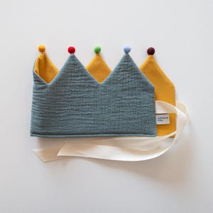 Könnte beinhalten: Eine Stoffkrone mit einer texturierten schieferblauen Außenseite und einer senfgelben Innenseite. Die Krone hat fünf Zacken, die jeweils mit einem kleinen Filz-Pompon in Gelb, Rot, Grün, Hellblau und Dunkelbraun verziert sind. Ein weißes Bindeband und ein Etikett mit der Aufschrift katharina kids sind angebracht.