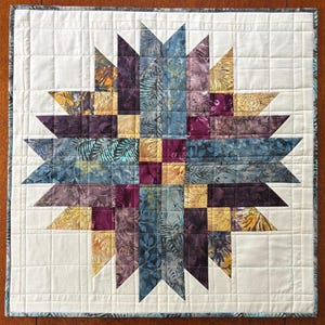 May include: A square fabric wall hanging featuring a central star pattern composed of batik fabric pieces in shades of purple, blue, and gold. The star is set against a cream-colored background with a grid-like quilted texture and a dark border.