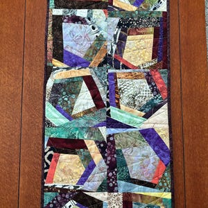 May include: A colorful patchwork table runner with geometric shapes in shades of purple, brown, green, and cream. The runner is made of fabric scraps sewn together in a random pattern.