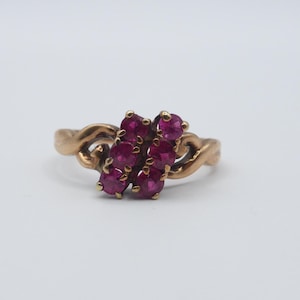 May include: A gold-toned ring featuring a cluster of seven round, deep red gemstones. The ring's band has a twisted design, and the gemstones are set in a floral-like arrangement. The ring is set against a white background.