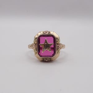 May include: A gold ring featuring a rectangular, deep pink gemstone with a gold star in the center. The ring has an ornate, floral design around the gemstone and on the band, showcasing a vintage aesthetic.