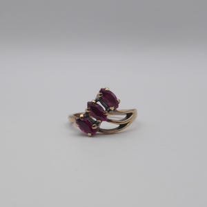 May include: A gold-toned ring featuring three marquise-cut, deep red gemstones. The ring has a split shank design, with the gemstones set in a cluster. The ring is set against a plain, light background.