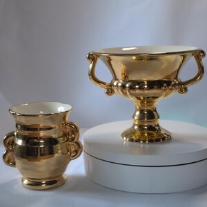May include: A pair of metallic gold ceramic vessels with a reflective finish. One is a wide, footed bowl with two curved handles, and the other is a smaller, cylindrical vase with two rounded handles on the sides and a cream-coloured interior.