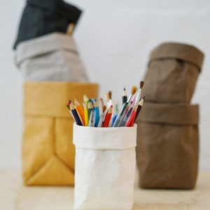 Könnte beinhalten: Eine weiße Papiertüte gefüllt mit bunten Bleistiften und Stiften, die auf einem Holztisch steht. Die Tüte ist von drei weiteren Papiertüten umgeben, einer schwarzen, einer grauen und einer braunen.