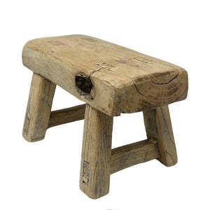 May include: A small, rectangular wooden stool with a light brown, weathered finish. The seat features a prominent natural knot hole and visible joinery details. Four sturdy, slightly angled legs are connected by horizontal support bars.