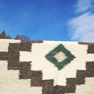 Puede incluir: Una alfombra tejida blanca y marr&oacute;n con un patr&oacute;n de diamante verde. La alfombra est&aacute; colgada contra un cielo azul con nubes blancas.