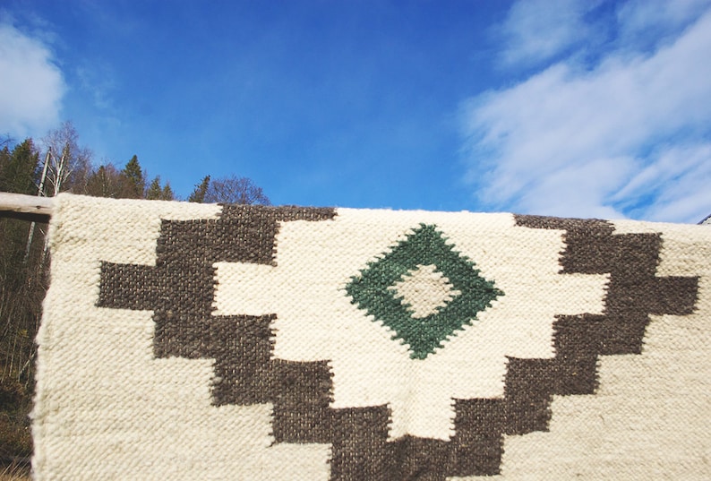 Puede incluir: Una alfombra tejida blanca y marr&oacute;n con un patr&oacute;n de diamante verde. La alfombra est&aacute; colgada contra un cielo azul con nubes blancas.