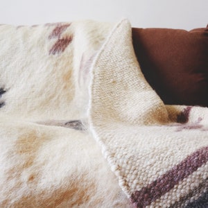 May include: A white and brown woven throw blanket with a fuzzy texture is draped over a brown couch. The blanket has a geometric pattern with stripes and a fringe edge.