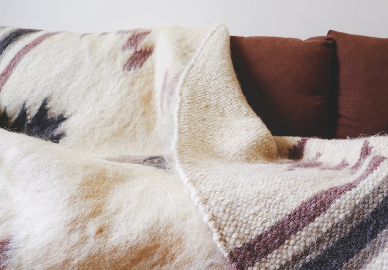 May include: A white and brown woven throw blanket with a fuzzy texture is draped over a brown couch. The blanket has a geometric pattern with stripes and a fringe edge.