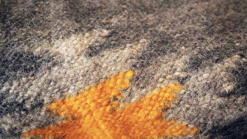 May include: Close-up of a gray and orange knitted fabric with a fuzzy texture. The fabric has a geometric pattern with a large orange section in the center.