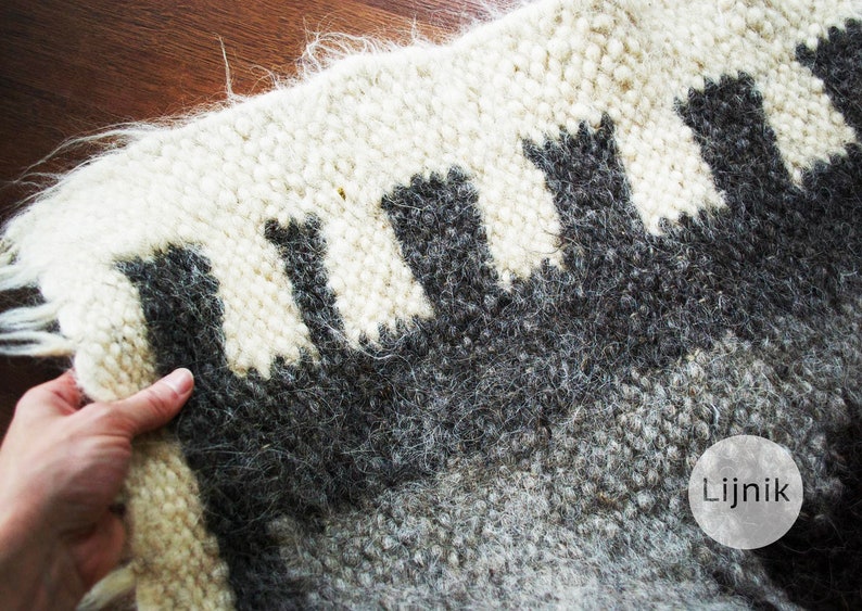 Puede incluir: Una alfombra de lana blanca y gris con un patr&oacute;n geom&eacute;trico de formas negras. La alfombra tiene una textura peluda y la palabra "Lijnik" est&aacute; impresa en un c&iacute;rculo en la esquina inferior derecha.