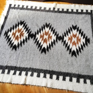 Puede incluir: Una alfombra tejida gris y blanca con un patr&oacute;n geom&eacute;trico de rombos marrones, negros y blancos. La alfombra tiene un borde de flecos.