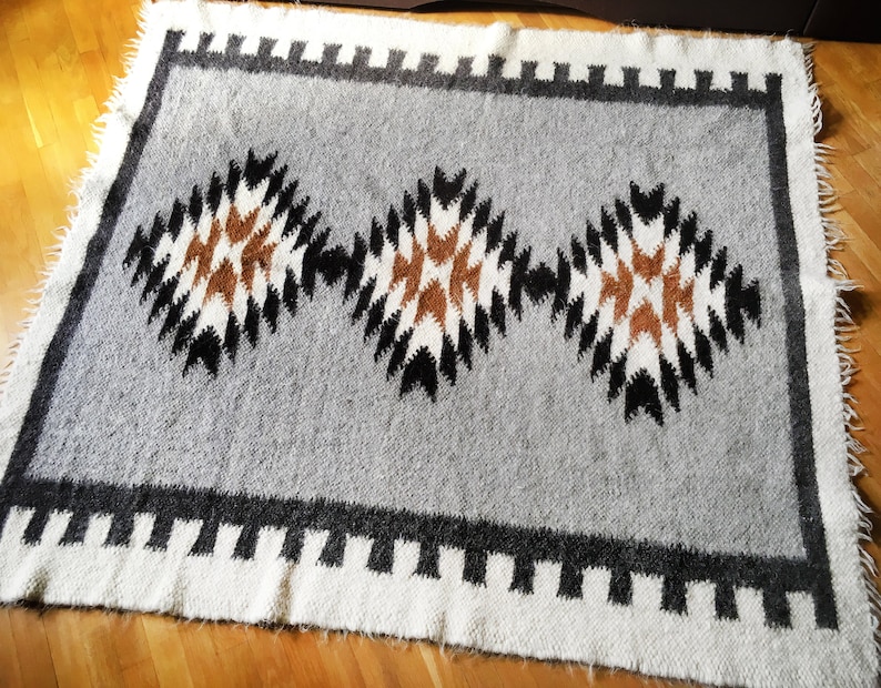 Puede incluir: Una alfombra tejida gris y blanca con un patr&oacute;n geom&eacute;trico de rombos marrones, negros y blancos. La alfombra tiene un borde de flecos.