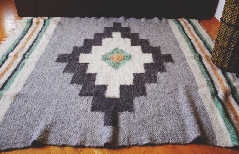 May include: A gray and white woven rug with a geometric pattern in black, green, and orange. The rug has a fringe border.